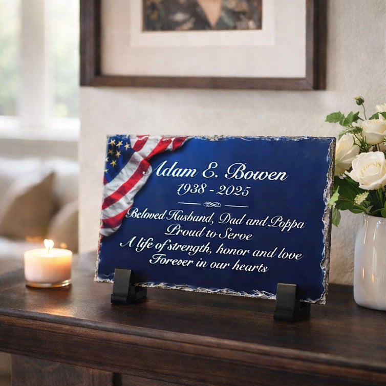 Memorial plaque with American flag design on a wooden surface, with a candle and flowers in the background.